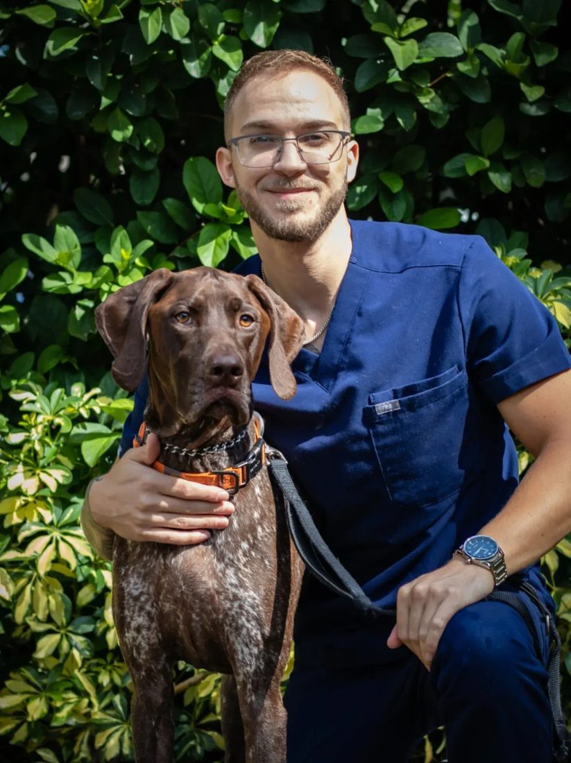 Man in blue scrubs kneeling with a brown dog outdoors