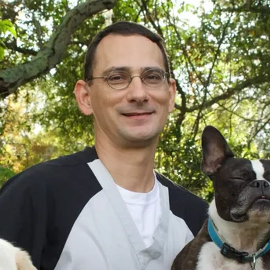 Man with glasses holding a French bulldog outdoors with trees